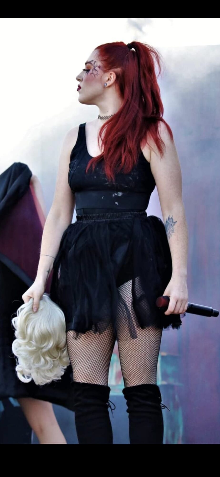 Woman with red hair in black dress holding a blonde wig, standing outdoors, side profile, sunglasses, tattoo on arm.