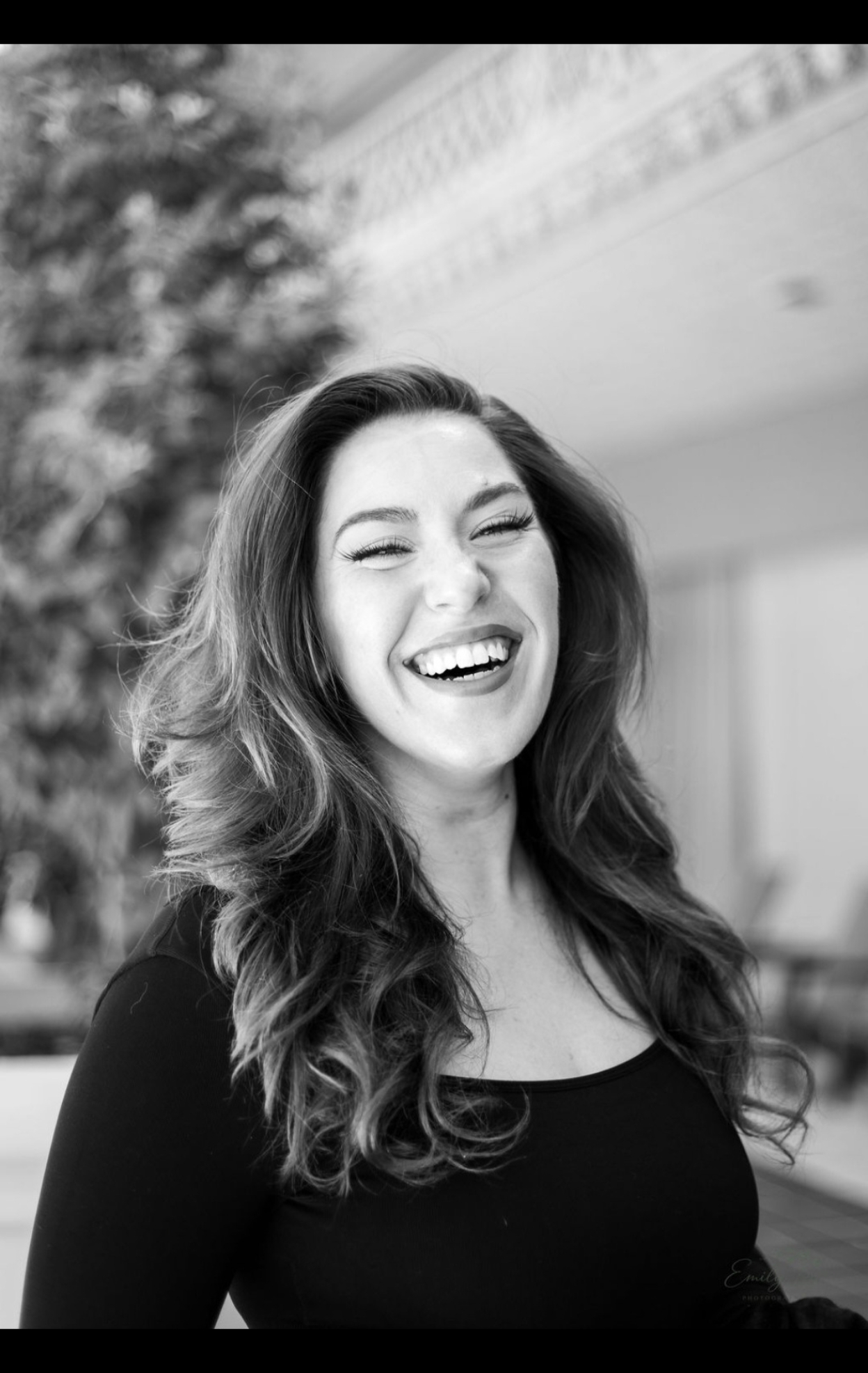 Woman with wavy hair laughing outdoors, background blurred, wearing a black top.