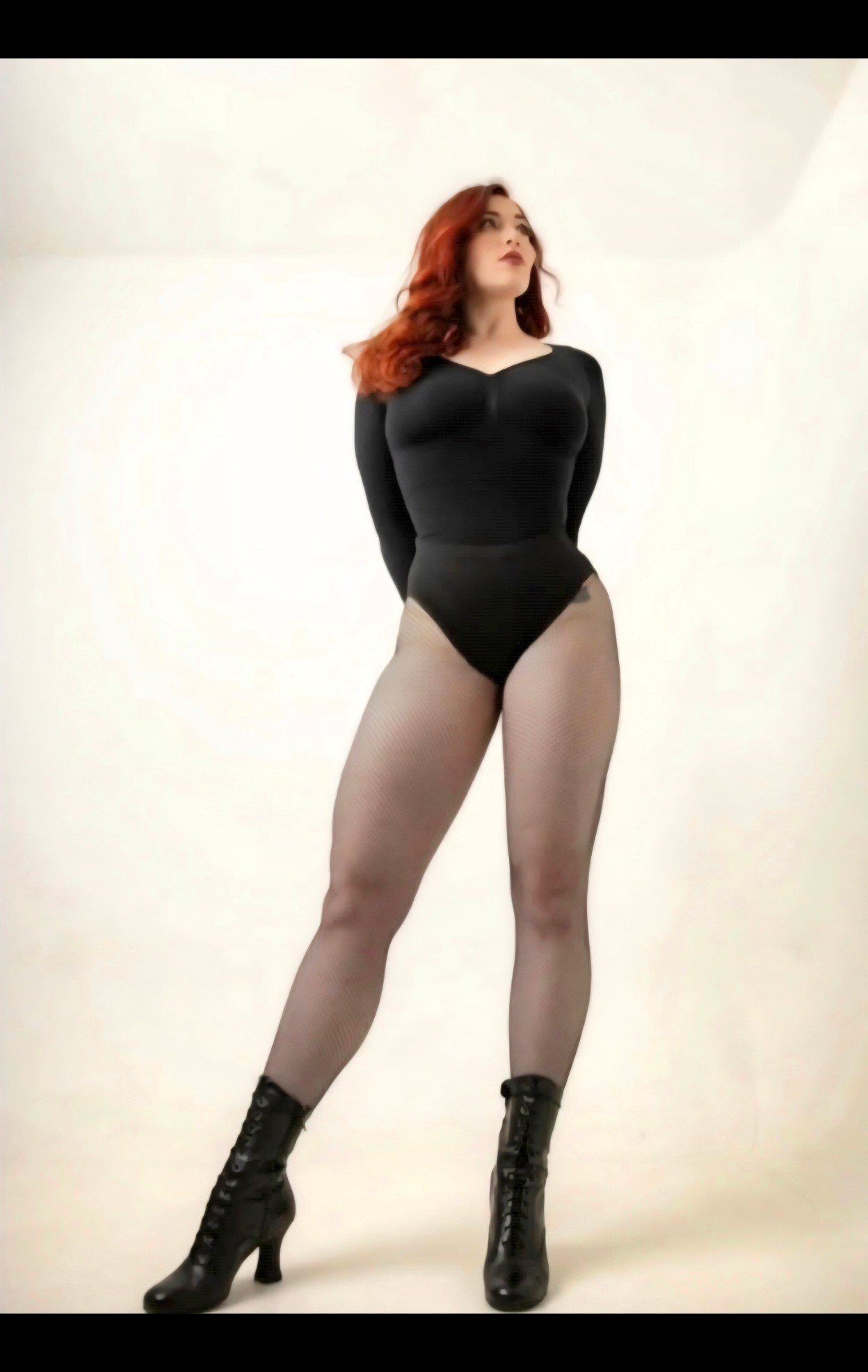 Woman with red hair in black leotard and high-heeled boots standing against a plain white background.