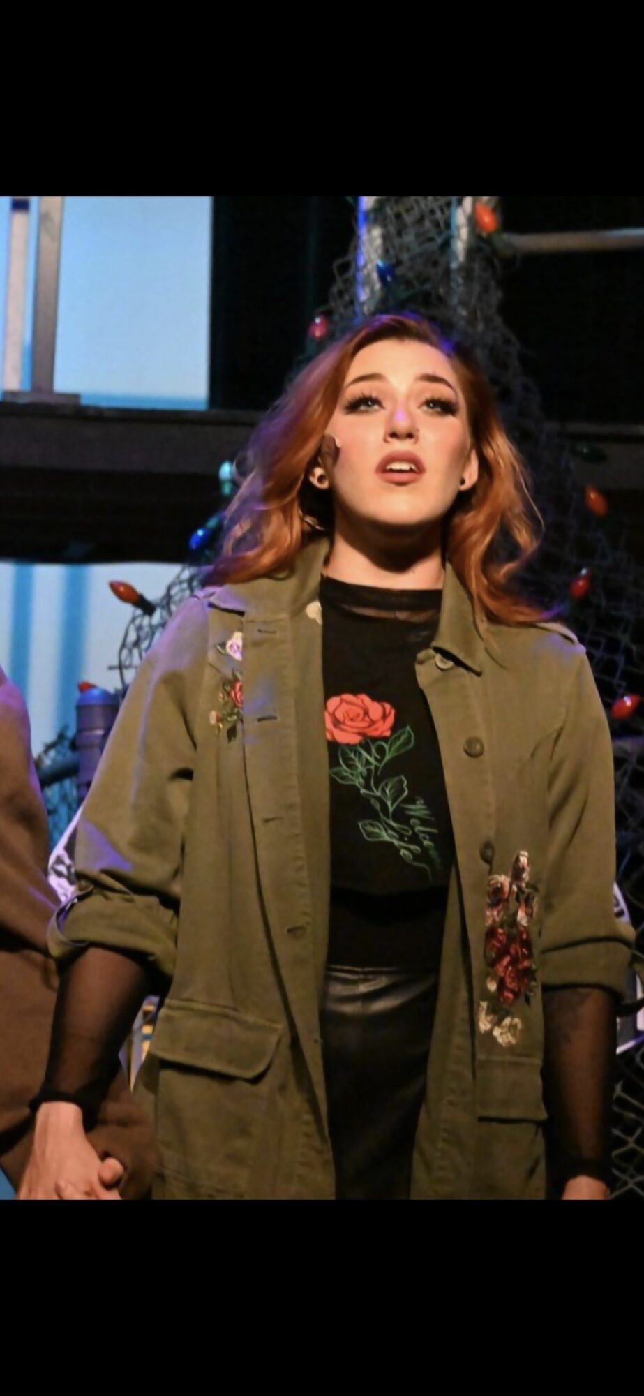Young woman with wavy hair wearing a green jacket and black shirt with floral design, standing indoors.