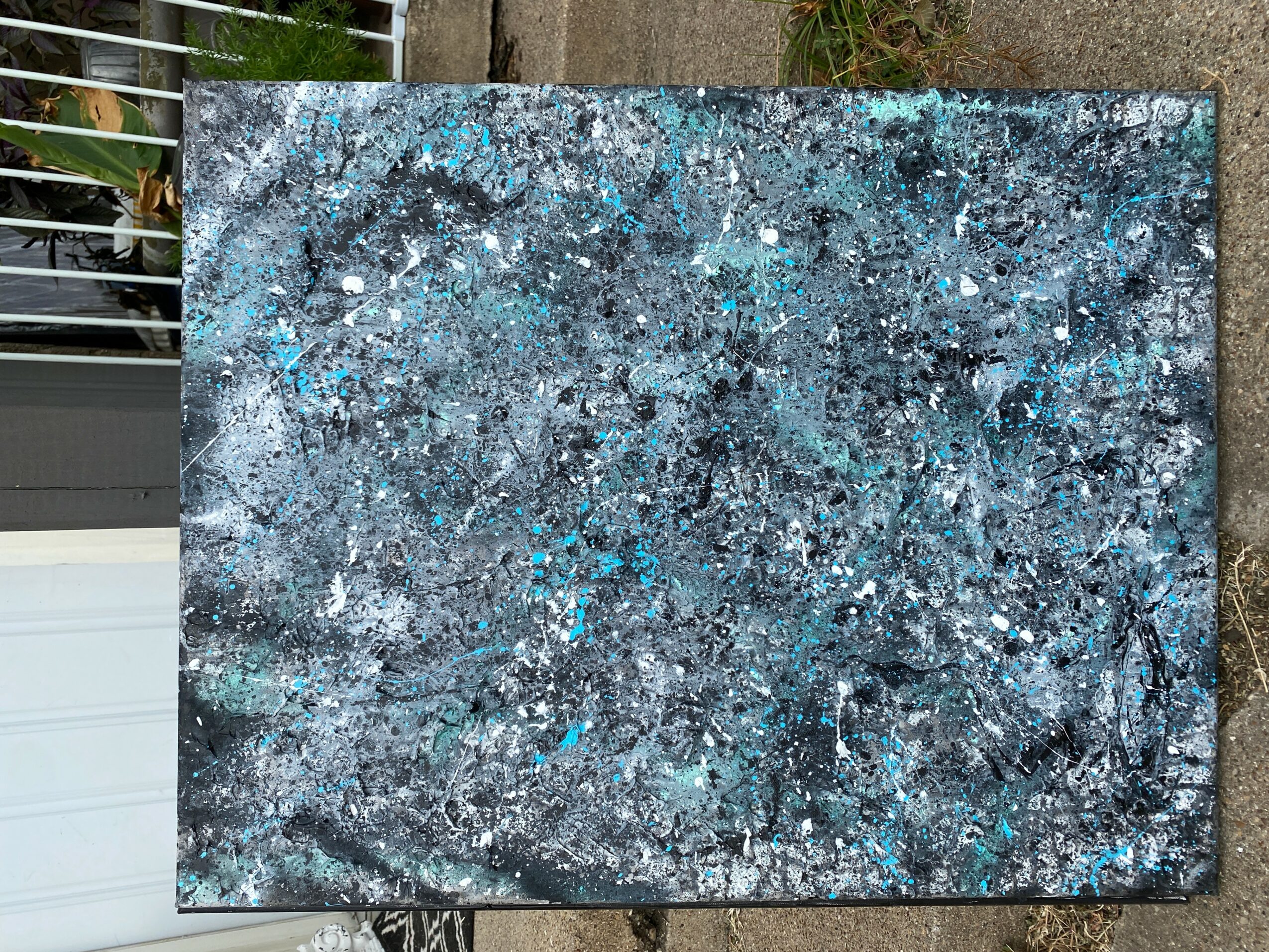 Abstract painting with blue, black, and white splatters on canvas, leaning against a wall outdoors.