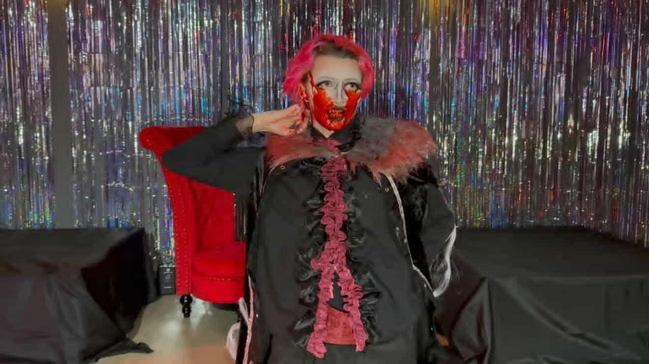 Person with red hair and face mask, wearing a dark coat with pink ruffles, sitting in front of a colorful backdrop.