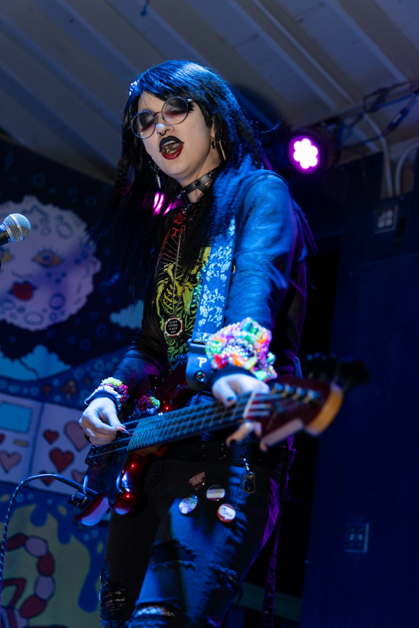 Person with long dark hair and glasses playing electric guitar on stage, wearing a blue jacket and colorful accessories.