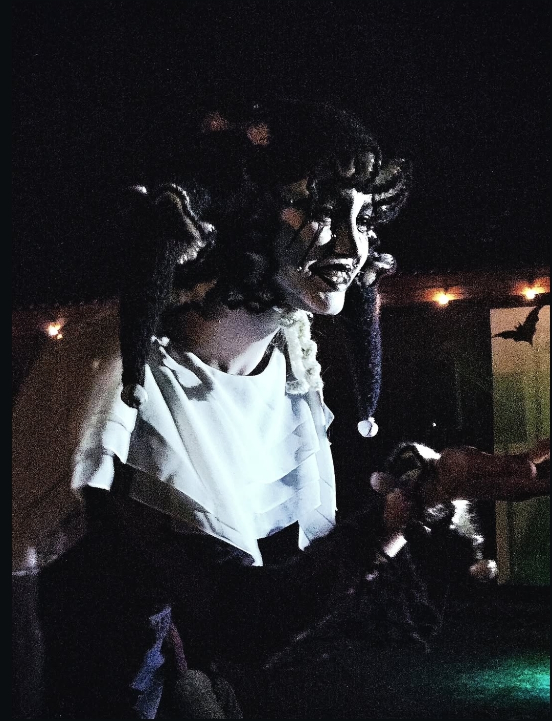 Person with dark curly hair and makeup, wearing a white top, in a dark environment with lights in the background.