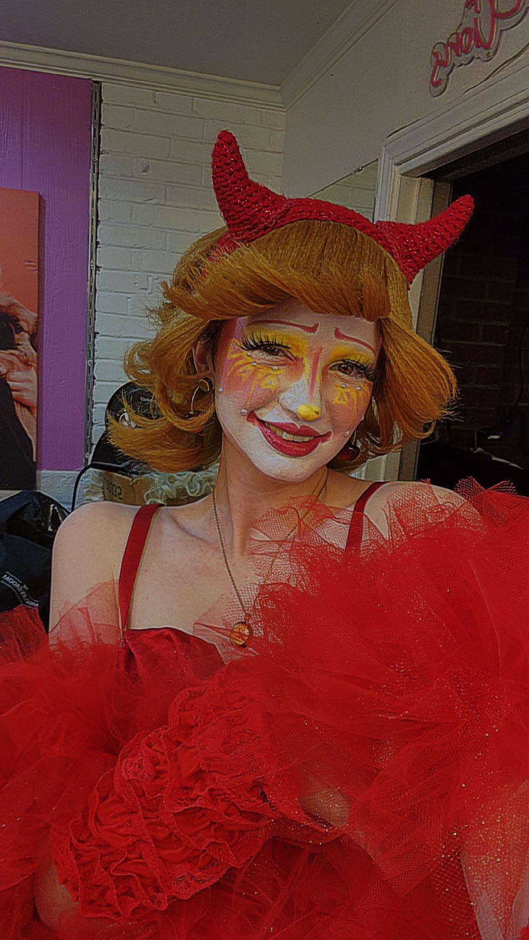 Person with red devil horns, orange hair, and colorful makeup, wearing a red costume with tulle, smiling.
