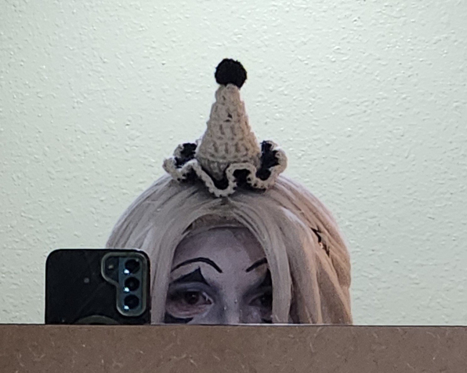 Person with light-colored hair wearing a small, knitted party hat with a pom-pom on top, reflected in a mirror.