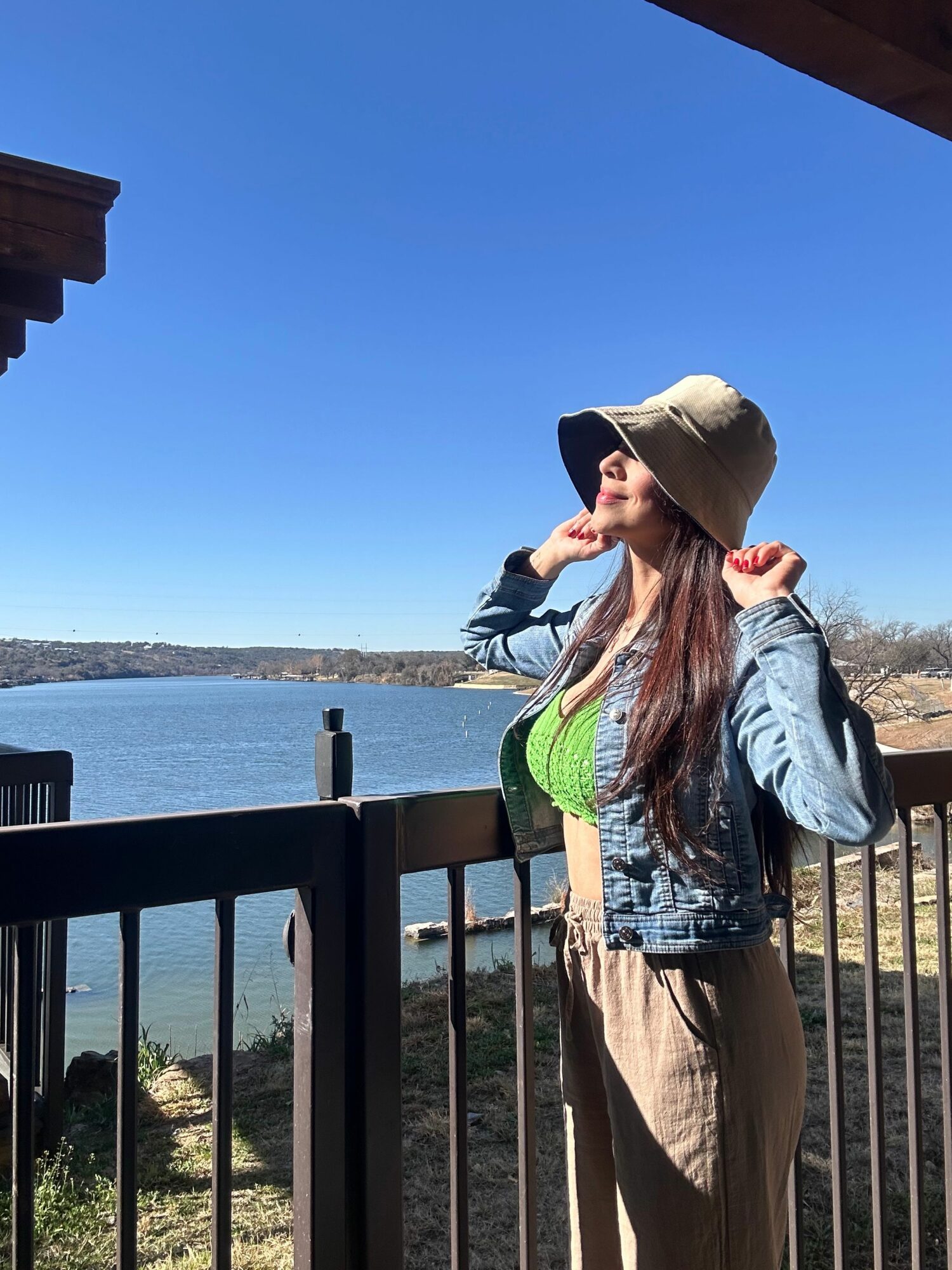 Woman wearing a hat and jacket stands on a balcony overlooking a body of water and landscape.