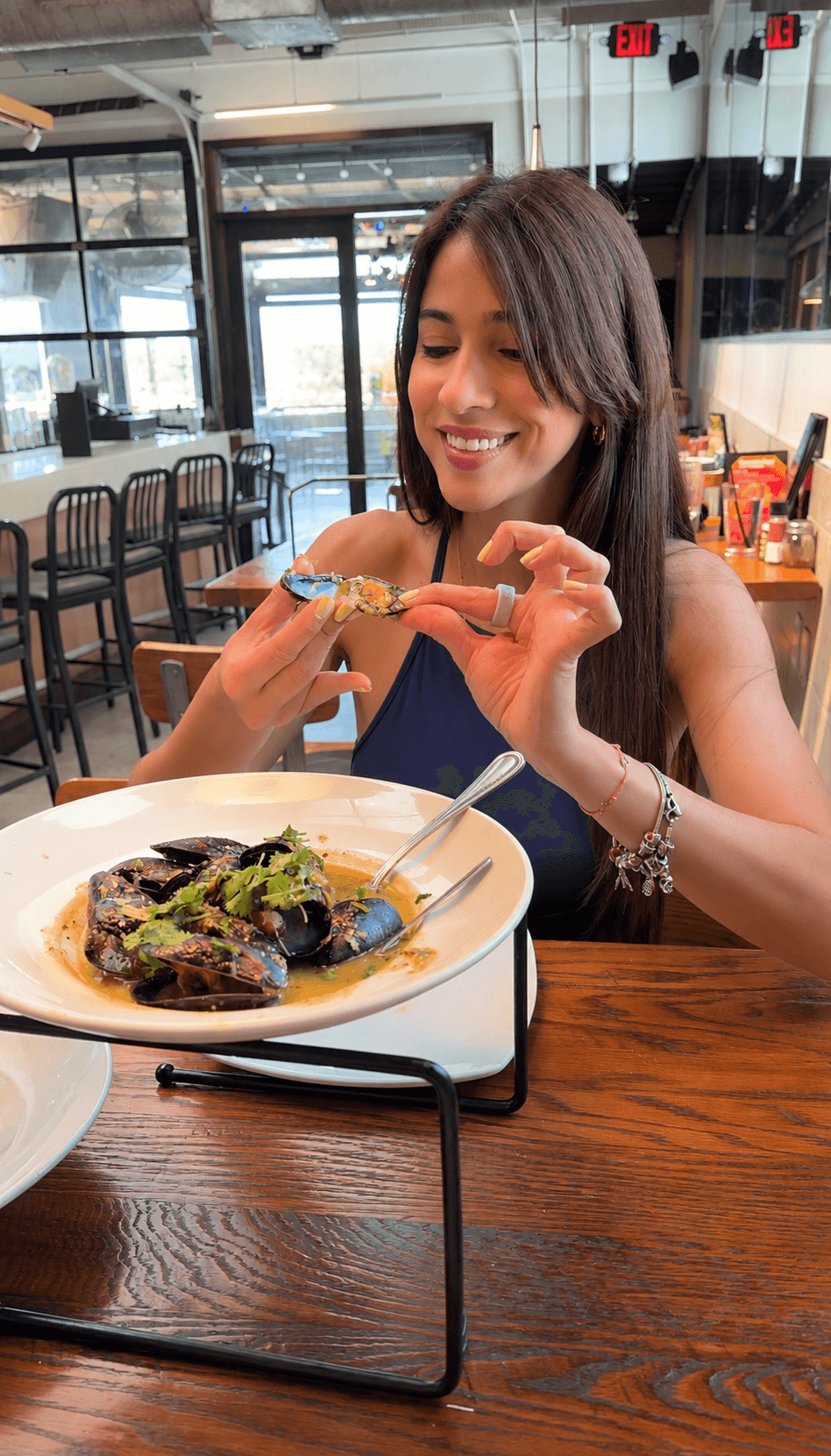 Woman smiling at a plate of food in a restaurant, holding a piece of bread, with empty chairs and windows in background.