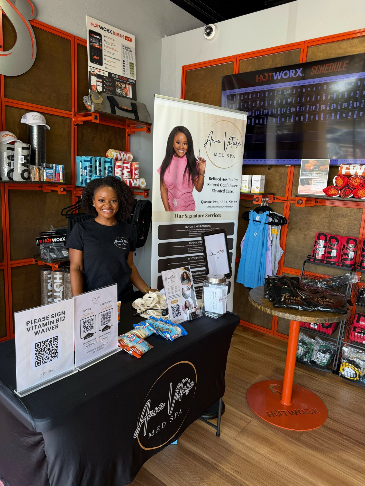Two women standing behind a table with promotional materials, in a store or event space with shelves and products in the background.