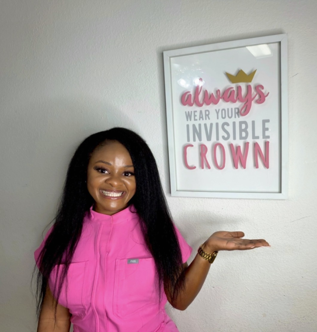 Smiling woman with long black hair wearing a pink top, gesturing towards a framed quote on the wall.