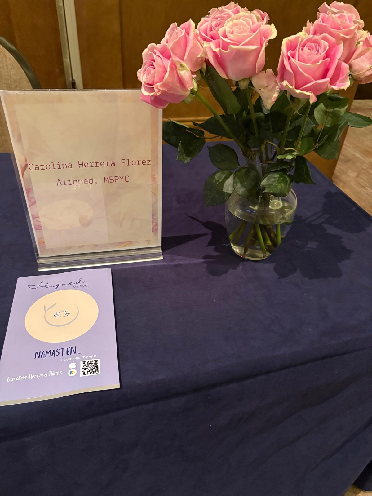 Pink roses in a glass vase on a dark table with a sign and a card nearby.