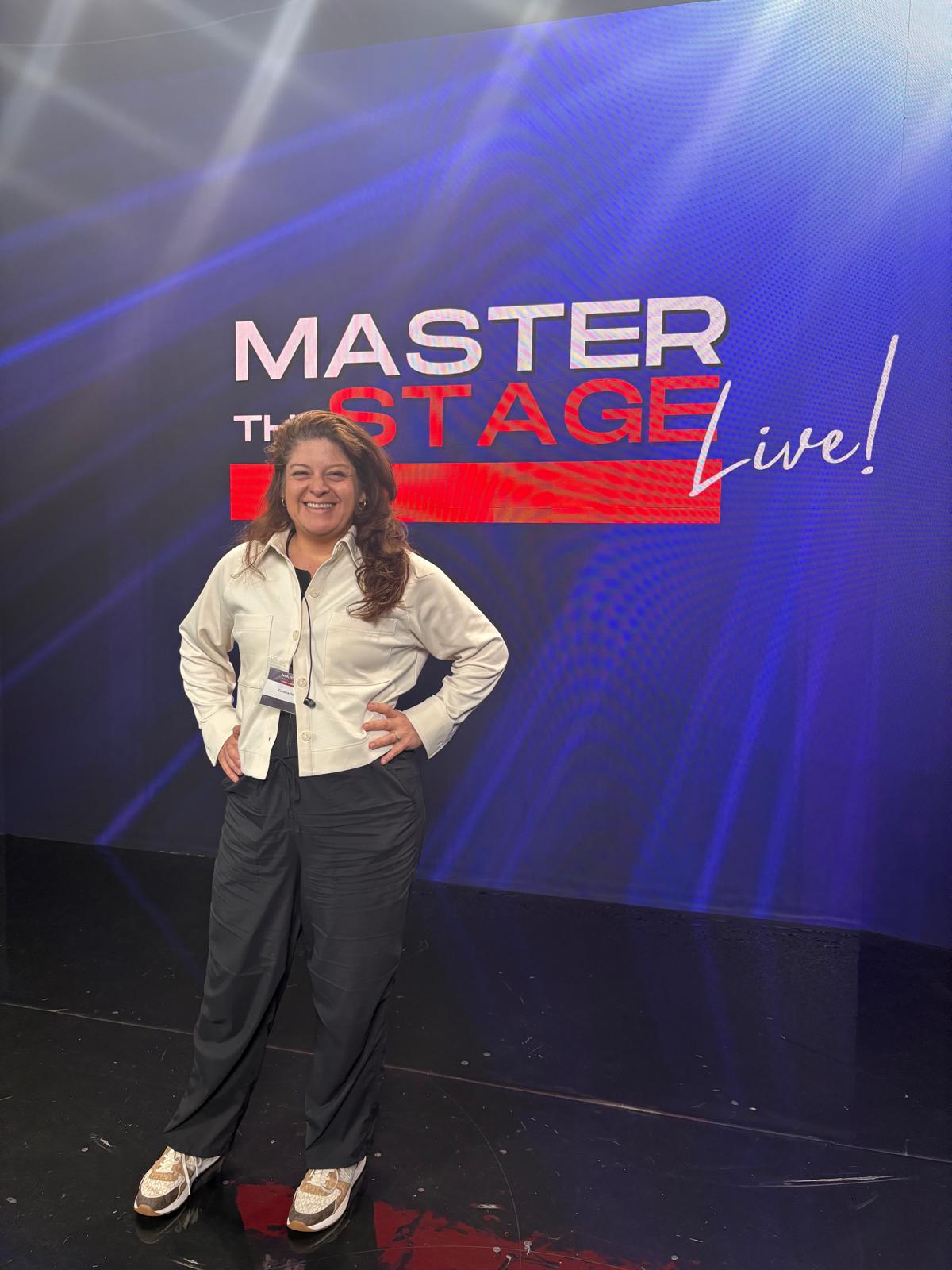 Woman standing in front of a stage backdrop with text, smiling, hands on hips, wearing a white jacket and black pants.