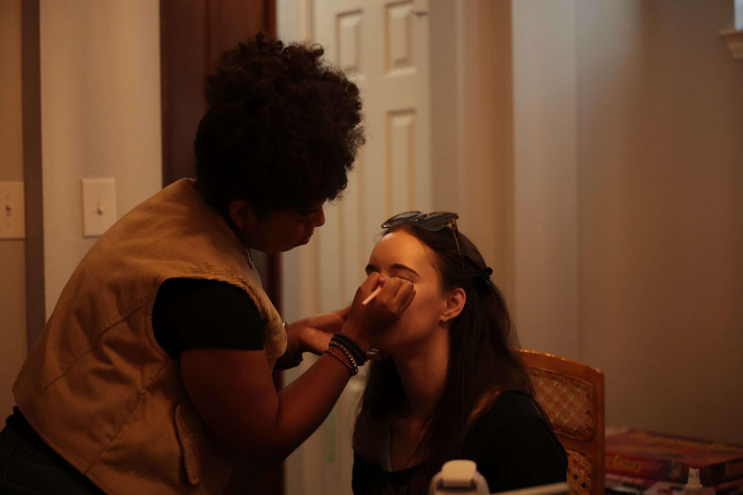 Person applying makeup to another person's face indoors, warm lighting, wooden door in background.