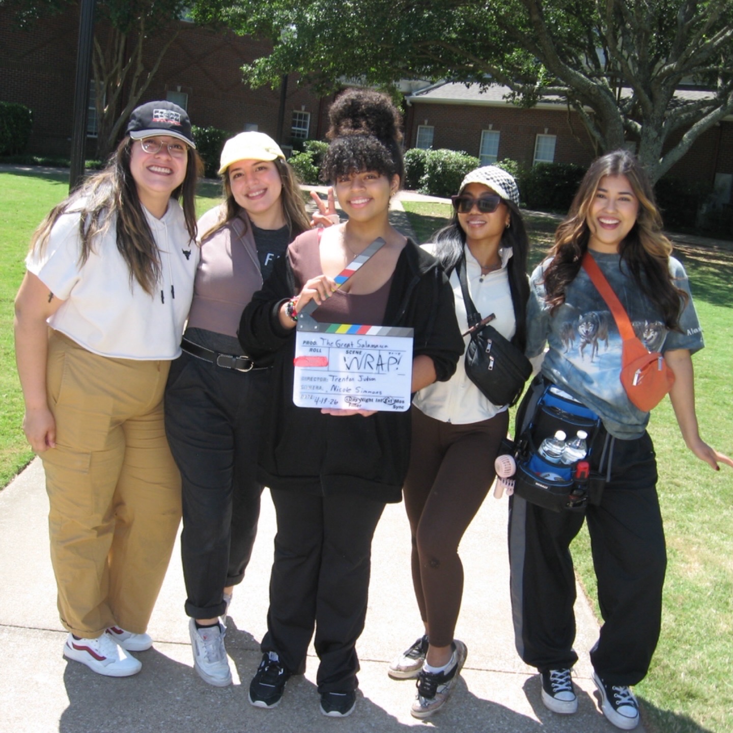 Group of five young women standing outdoors on grass, smiling, holding a sign, with trees and buildings in background.