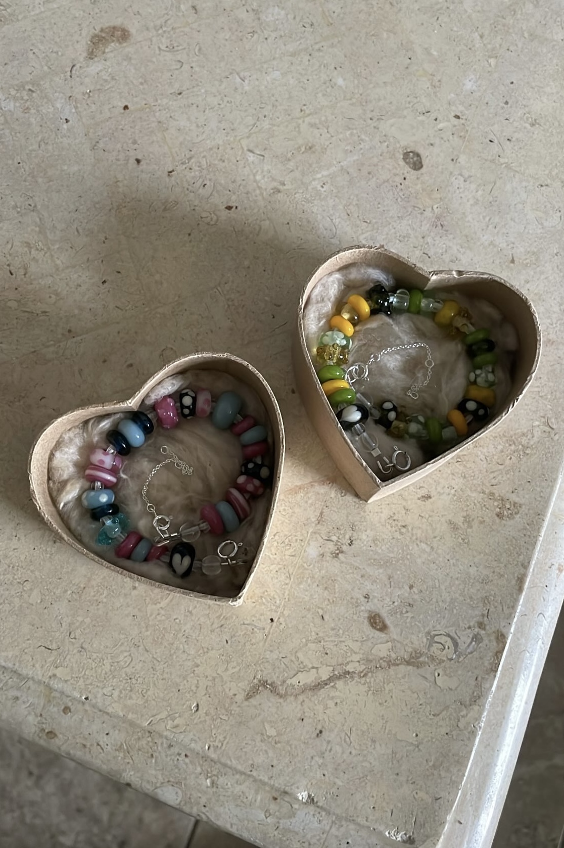 Two heart-shaped boxes with colorful beaded bracelets inside, placed on a light-colored surface.