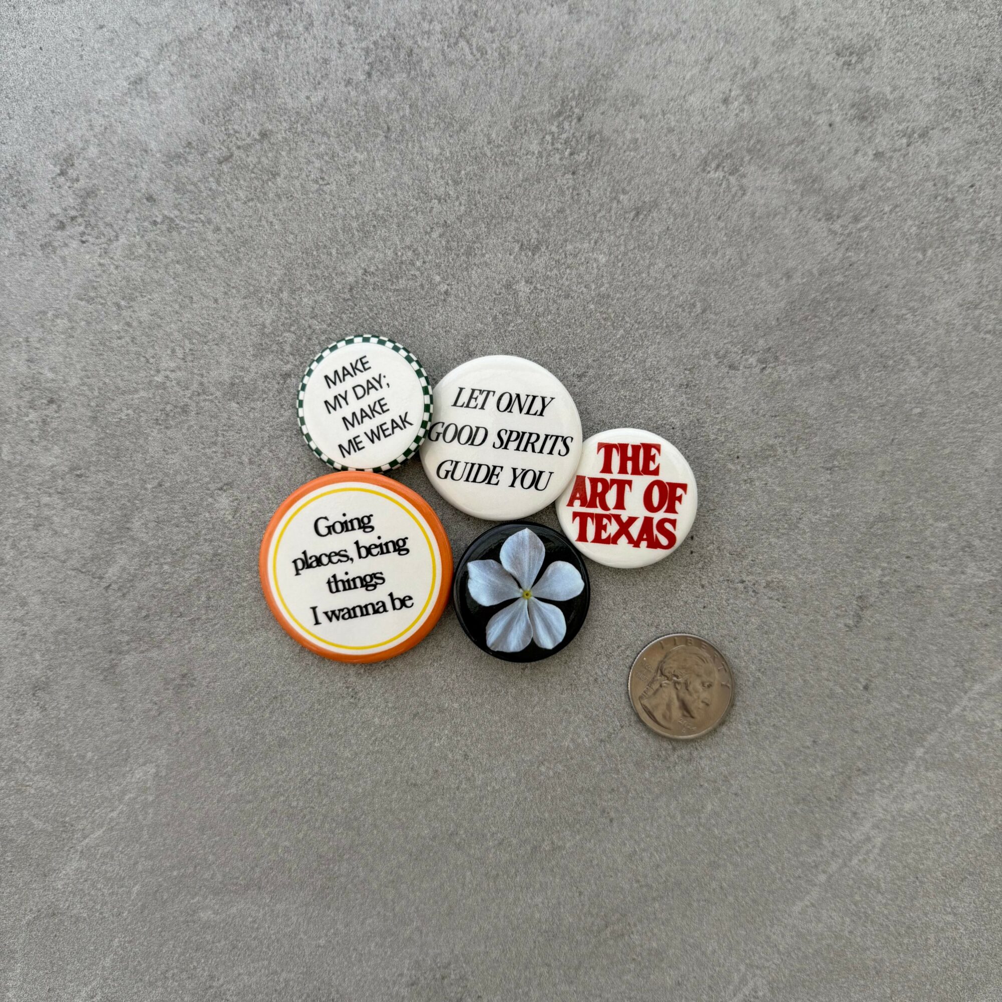 Six small round objects with text and a flower, and a coin on a gray surface.