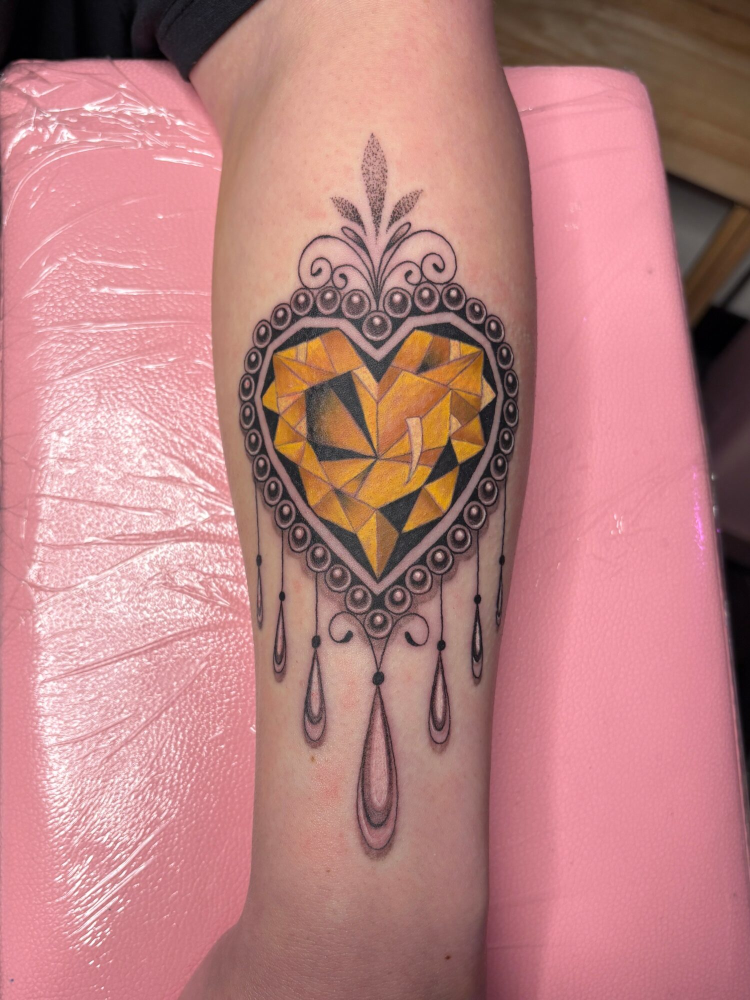 Tattoo of a geometric heart inside a decorative frame with hanging elements on a person's arm.
