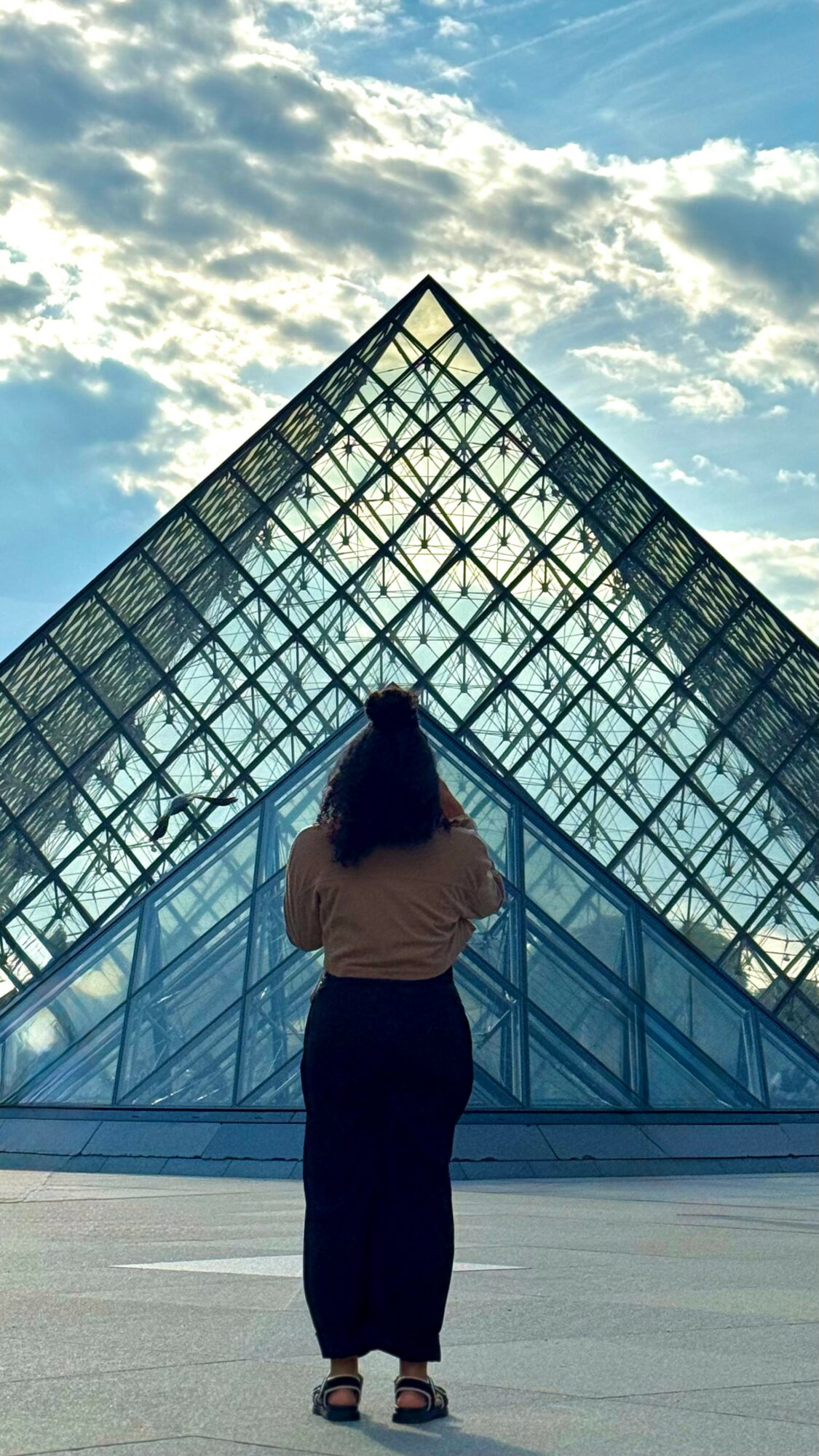 Person standing in front of a glass pyramid structure, looking up at the sky with clouds.