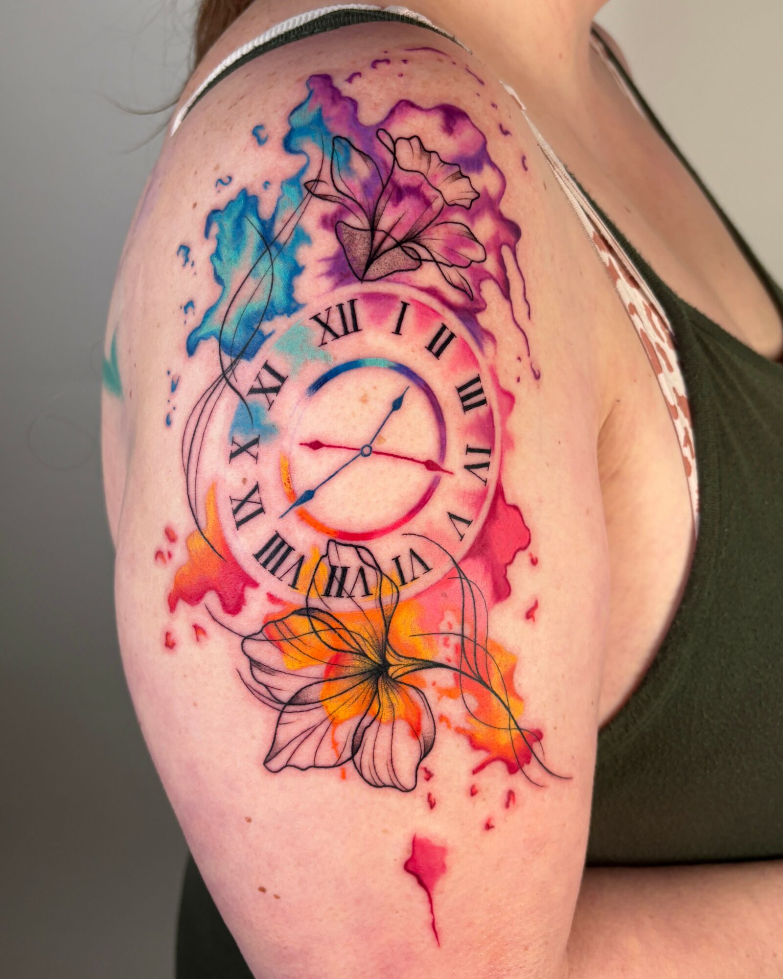 Colorful watercolor-style tattoo of a clock with Roman numerals, surrounded by flowers and abstract splashes on shoulder.