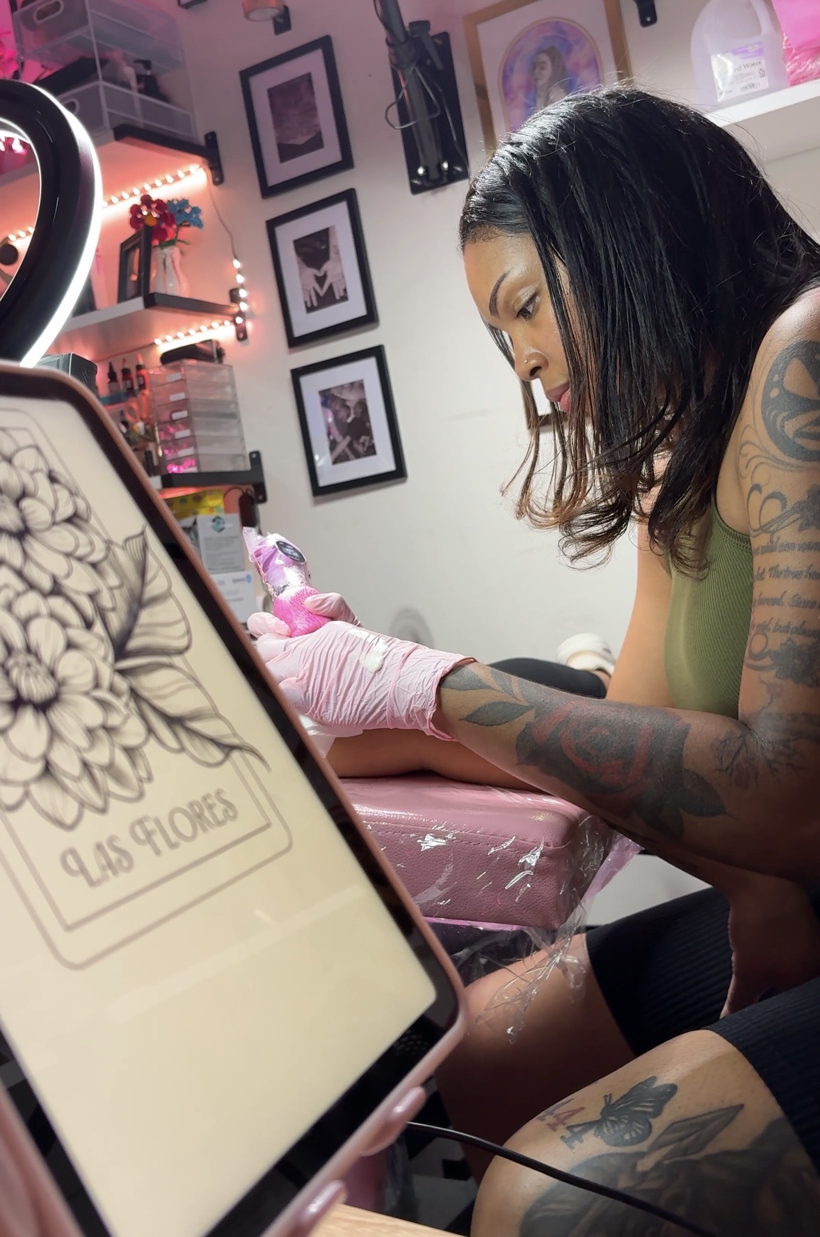 Tattooed woman getting a tattoo, focused on her arm, in a tattoo studio with framed pictures on the wall.