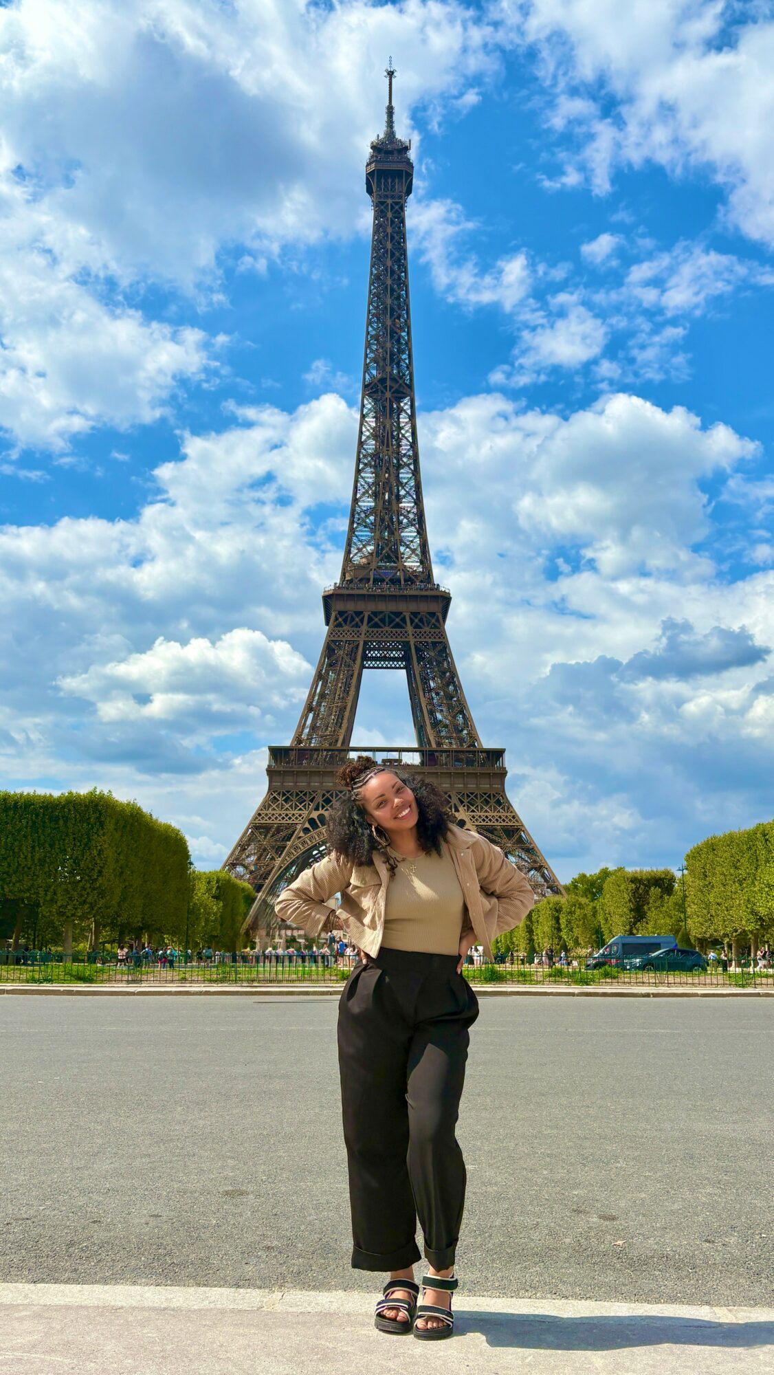 Woman standing in front of Eiffel Tower with blue sky and clouds, trees on sides, wearing beige top and black pants.