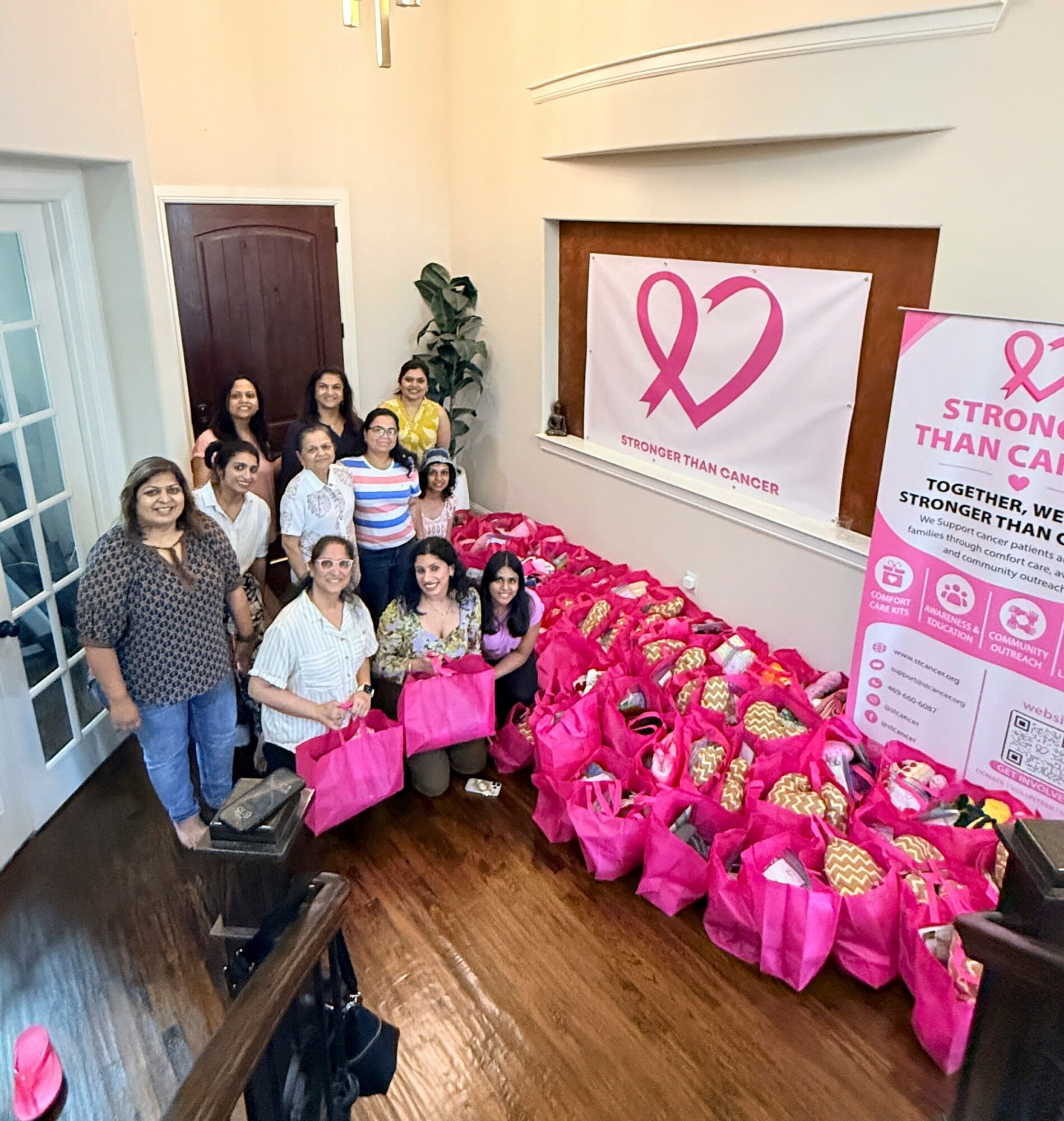 Group of women standing indoors with pink bags and breast cancer awareness banners, smiling at the camera.