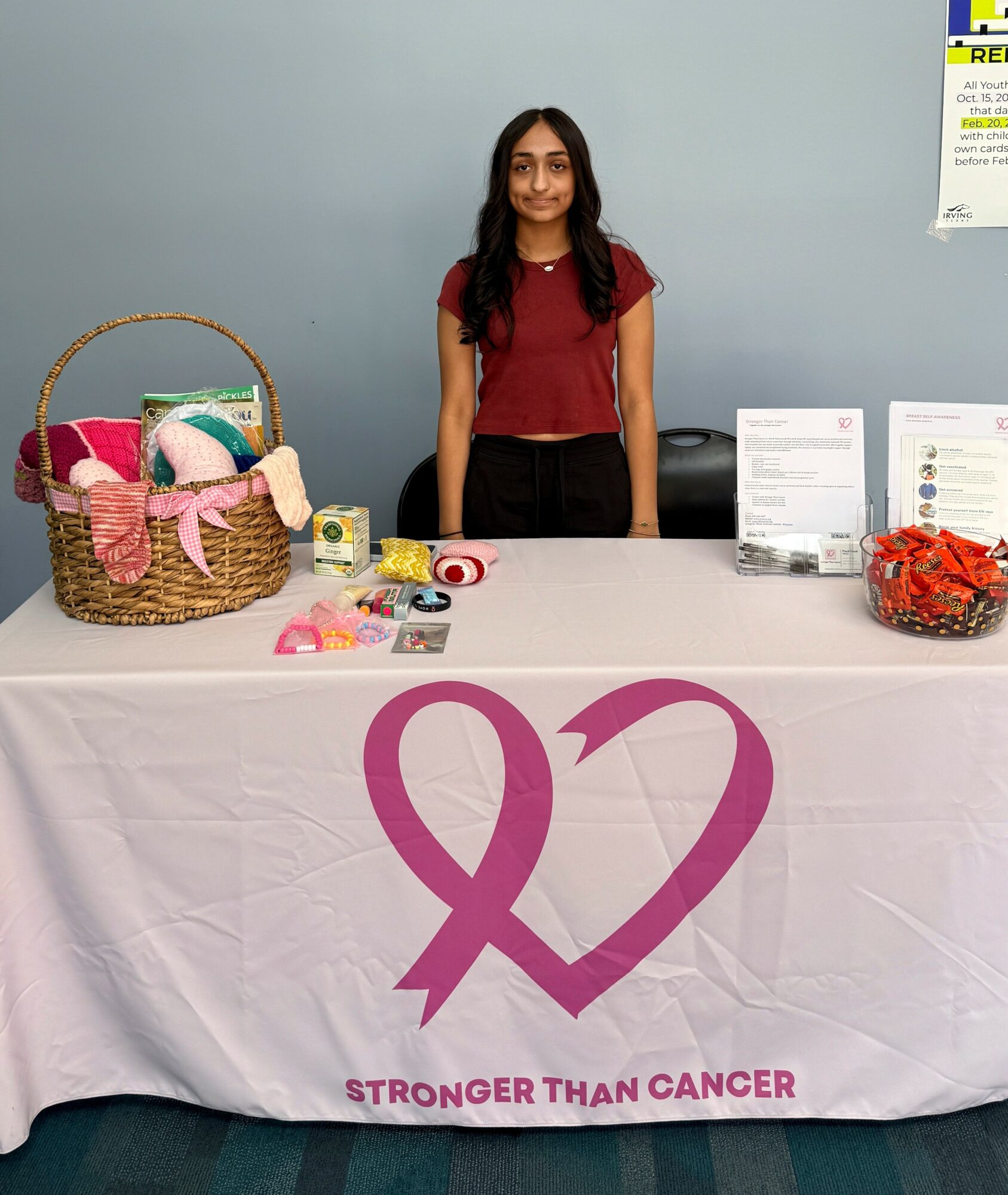 Young woman standing behind a table with a pink ribbon and cancer awareness items, against a light blue wall.