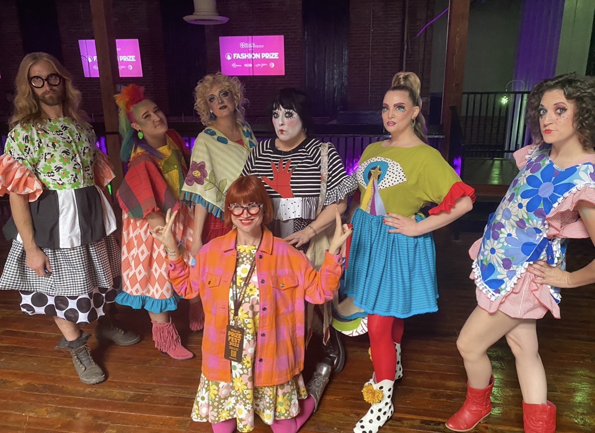 Group of seven women in colorful, eclectic costumes posing indoors with wooden floor and purple screens in background.