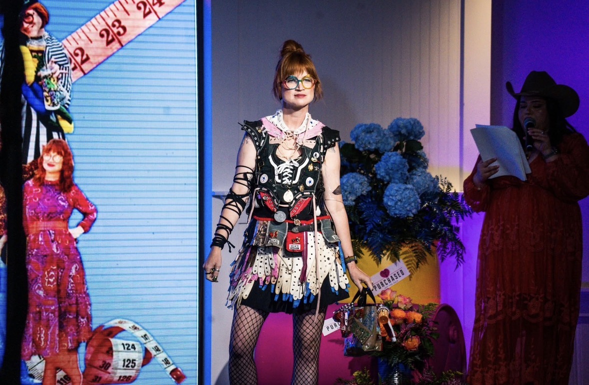 Woman in colorful costume with glasses, standing on stage with flowers and other performers, colorful background.