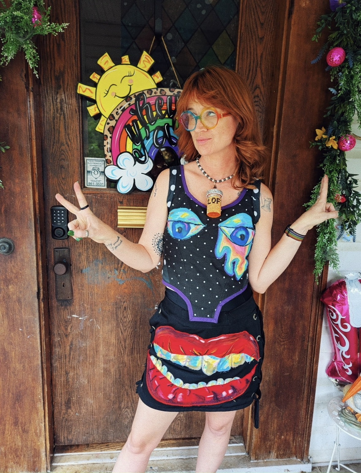 Woman with red hair and glasses stands with arms outstretched in front of a wooden door decorated with a sun and rainbow, wearing a colorful dress.