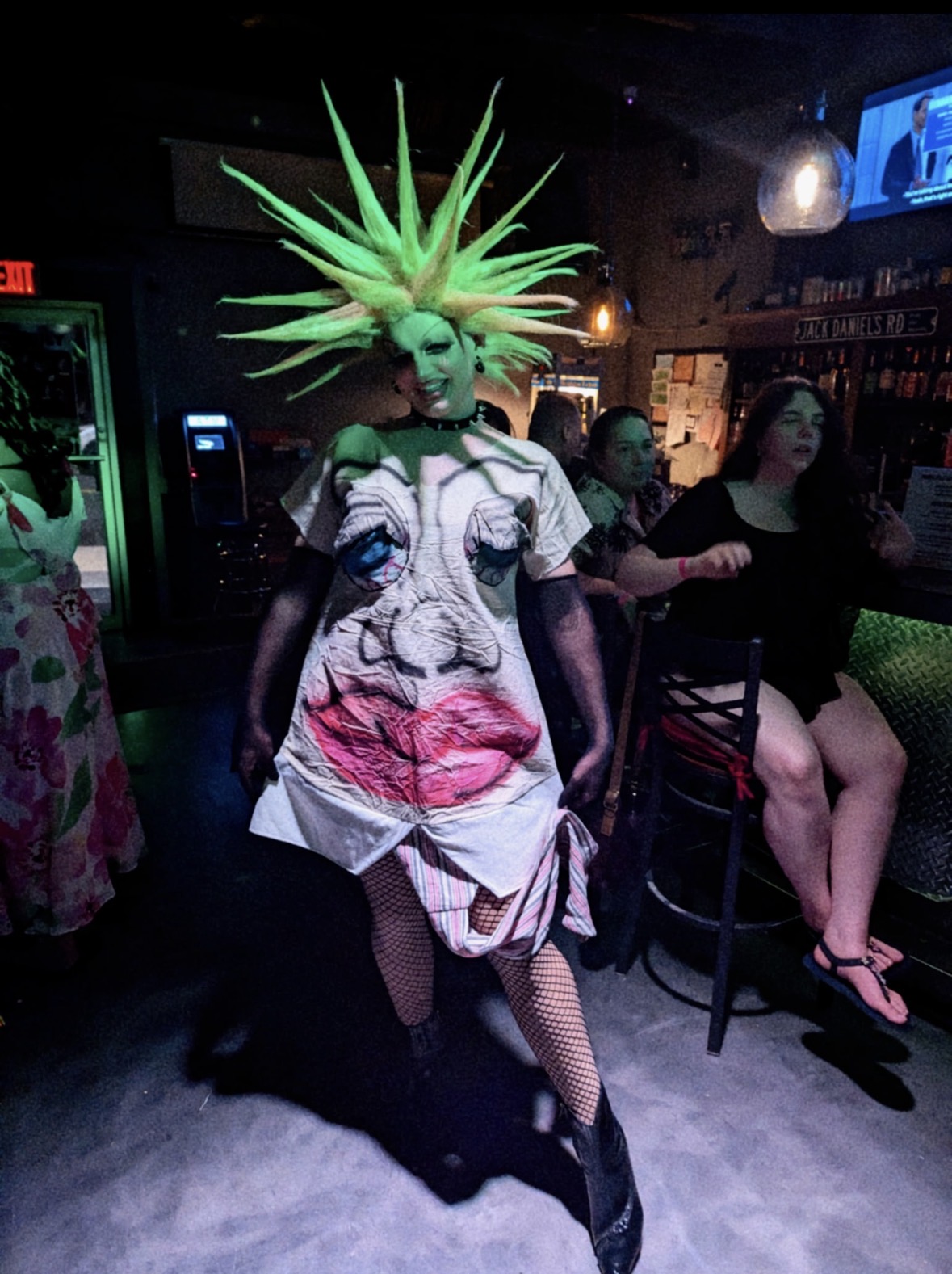 Person wearing a dress with a face and lips design, and a spiky green headpiece, in a dimly lit bar. Word count: 20