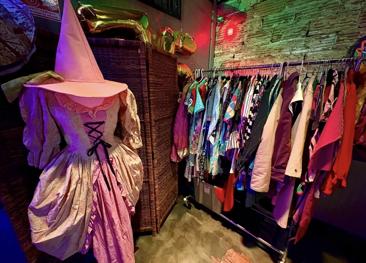 Clothing rack with colorful garments and a costume with a pointed hat in a room with brick wall and colorful lighting.
