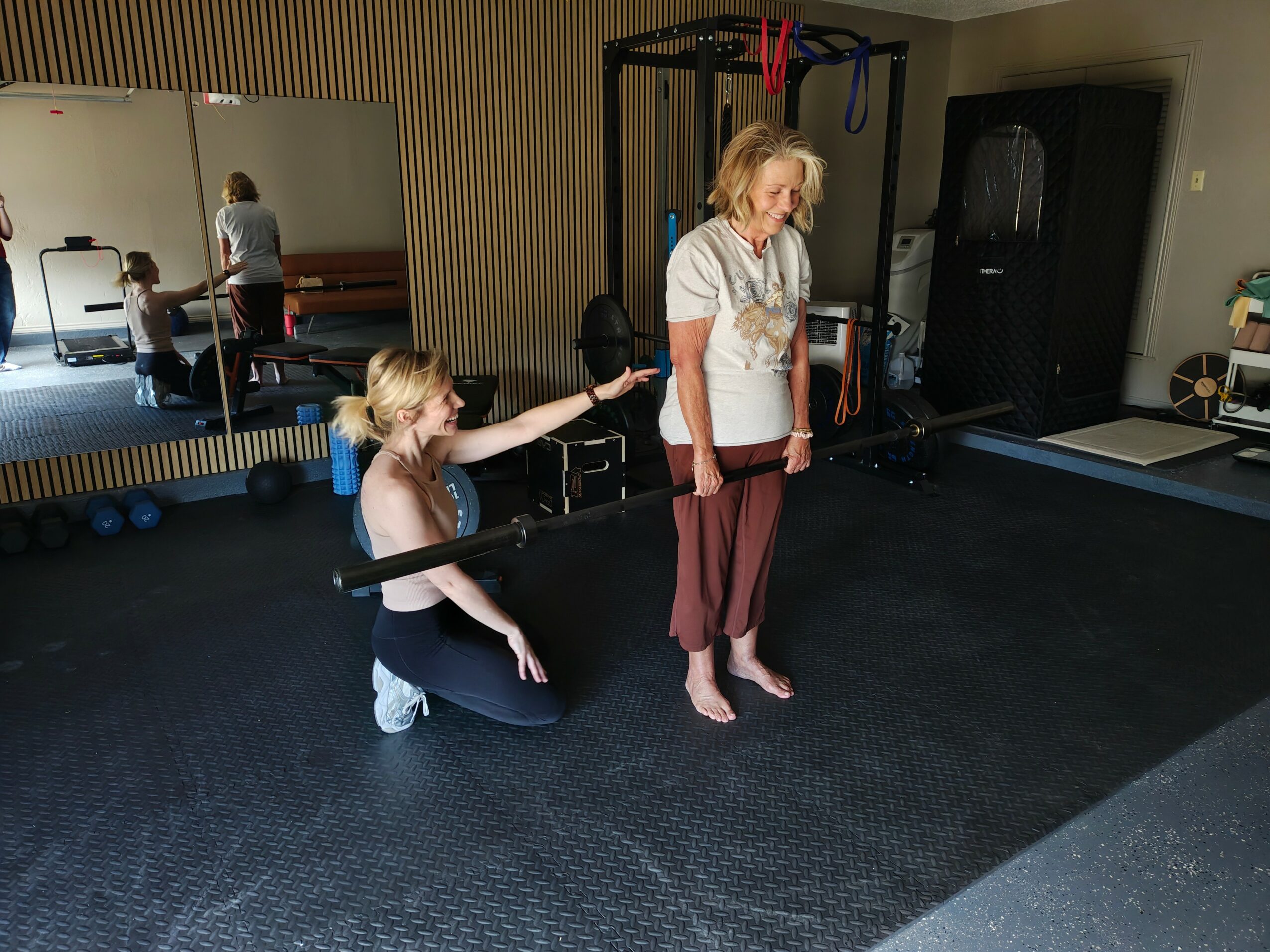 A woman standing barefoot in a gym with a young girl kneeling beside her, holding a barbell. Mirror reflects other people exercising.