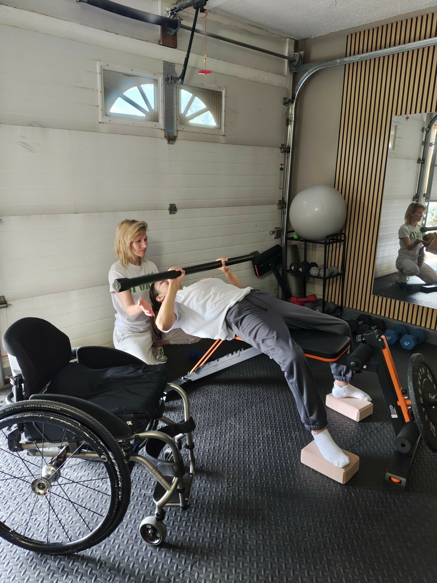 Person in a wheelchair working out with dumbbells in a gym, with exercise equipment and a mirror in the background.