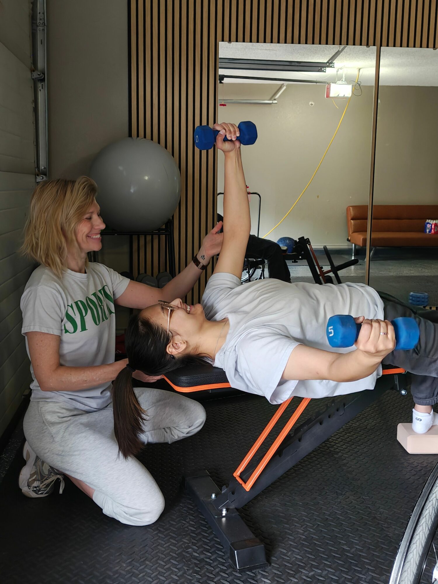 Two women exercising with dumbbells in a gym, one lying on a mat and the other sitting, with gym equipment around.