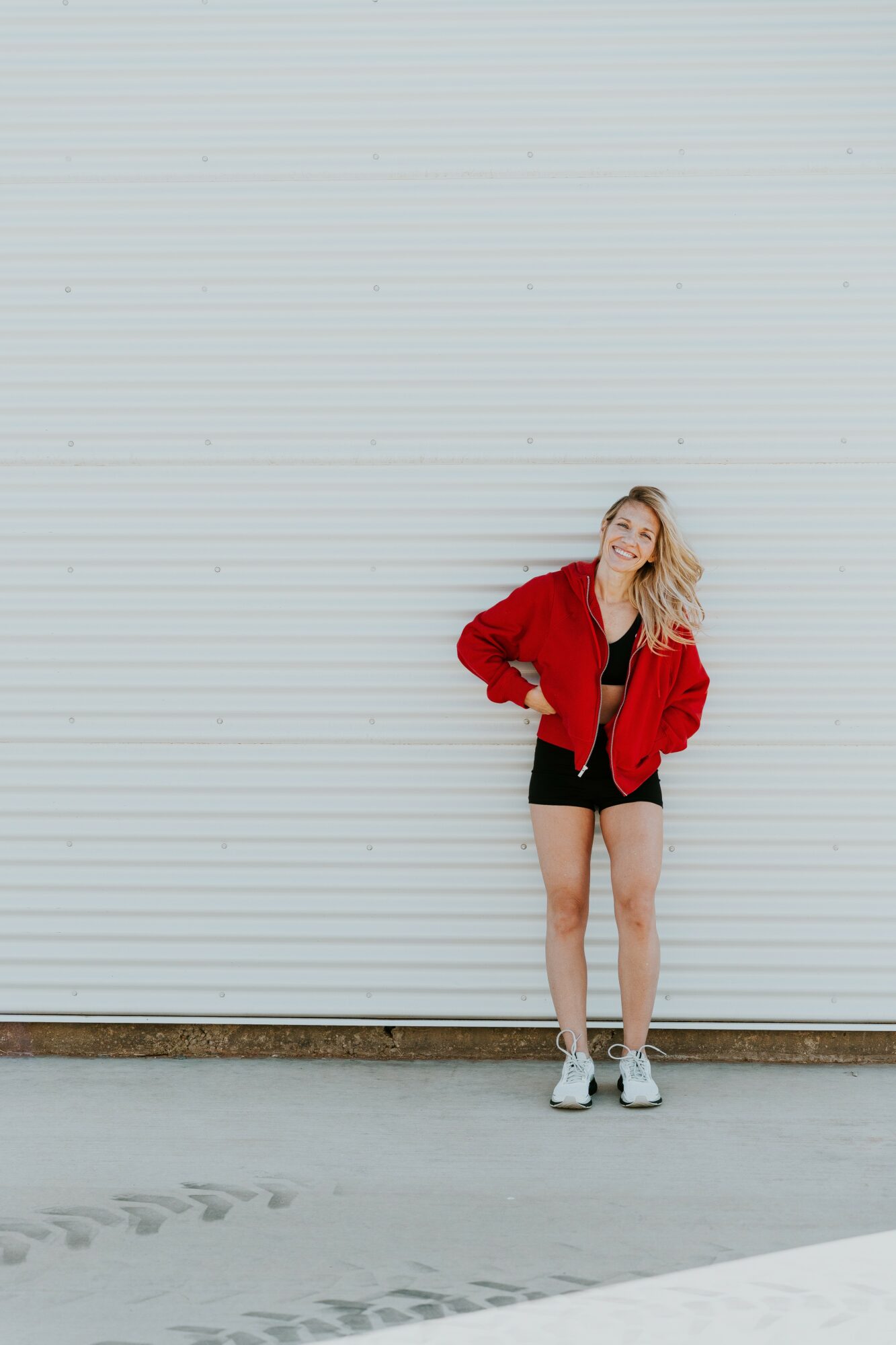 Young woman with blonde hair in a red jacket, black shorts, and sneakers standing against a large white wall.