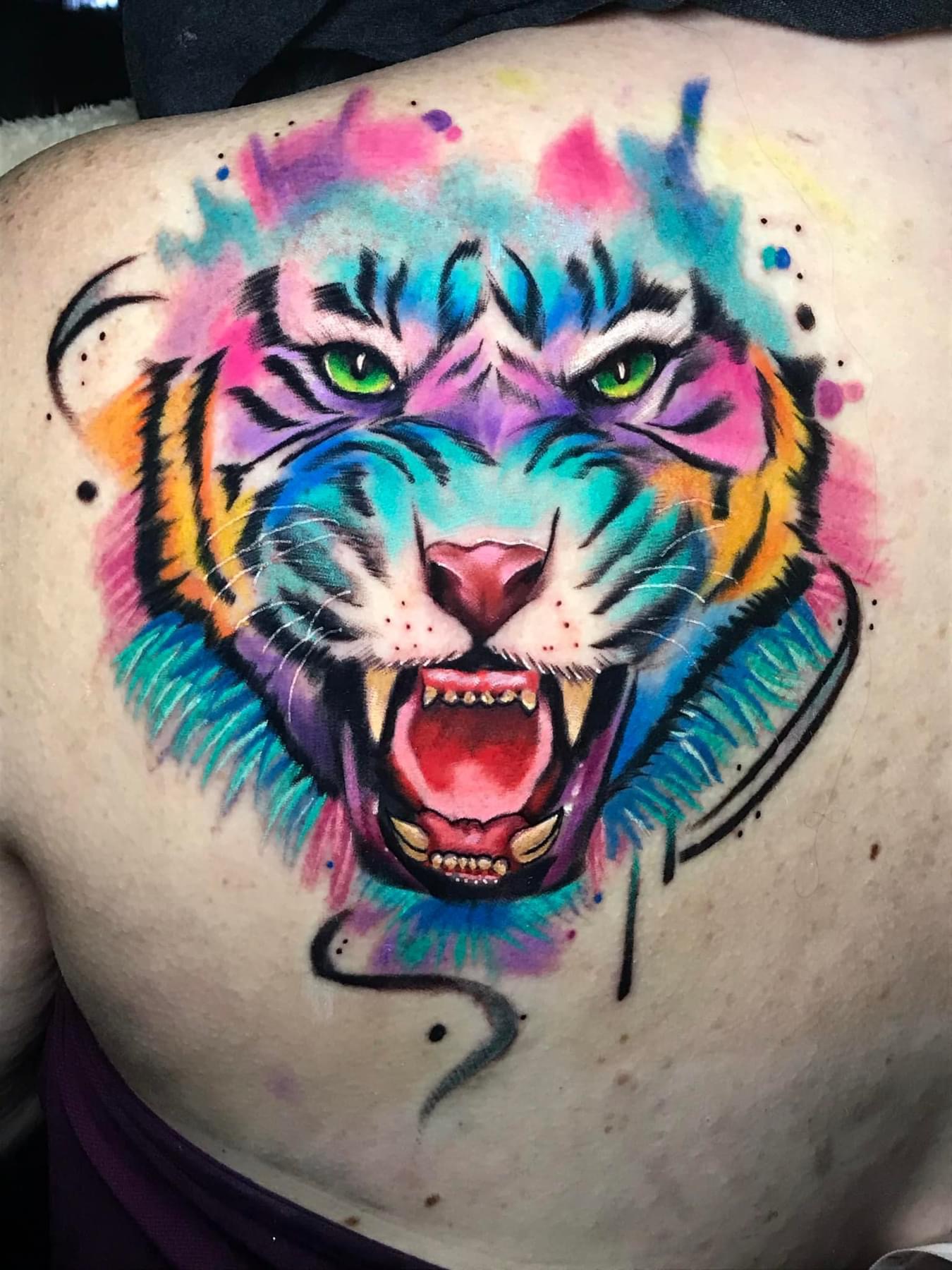 Colorful tiger face tattoo with green eyes, pink nose, and open mouth showing teeth, surrounded by vibrant splashes of color.