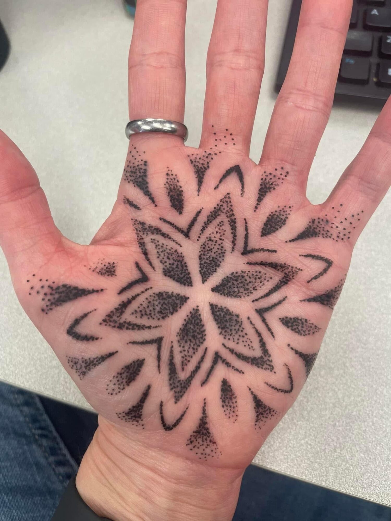 Hand with a floral tattoo design in black ink covering the palm and fingers, with a ring on the middle finger.