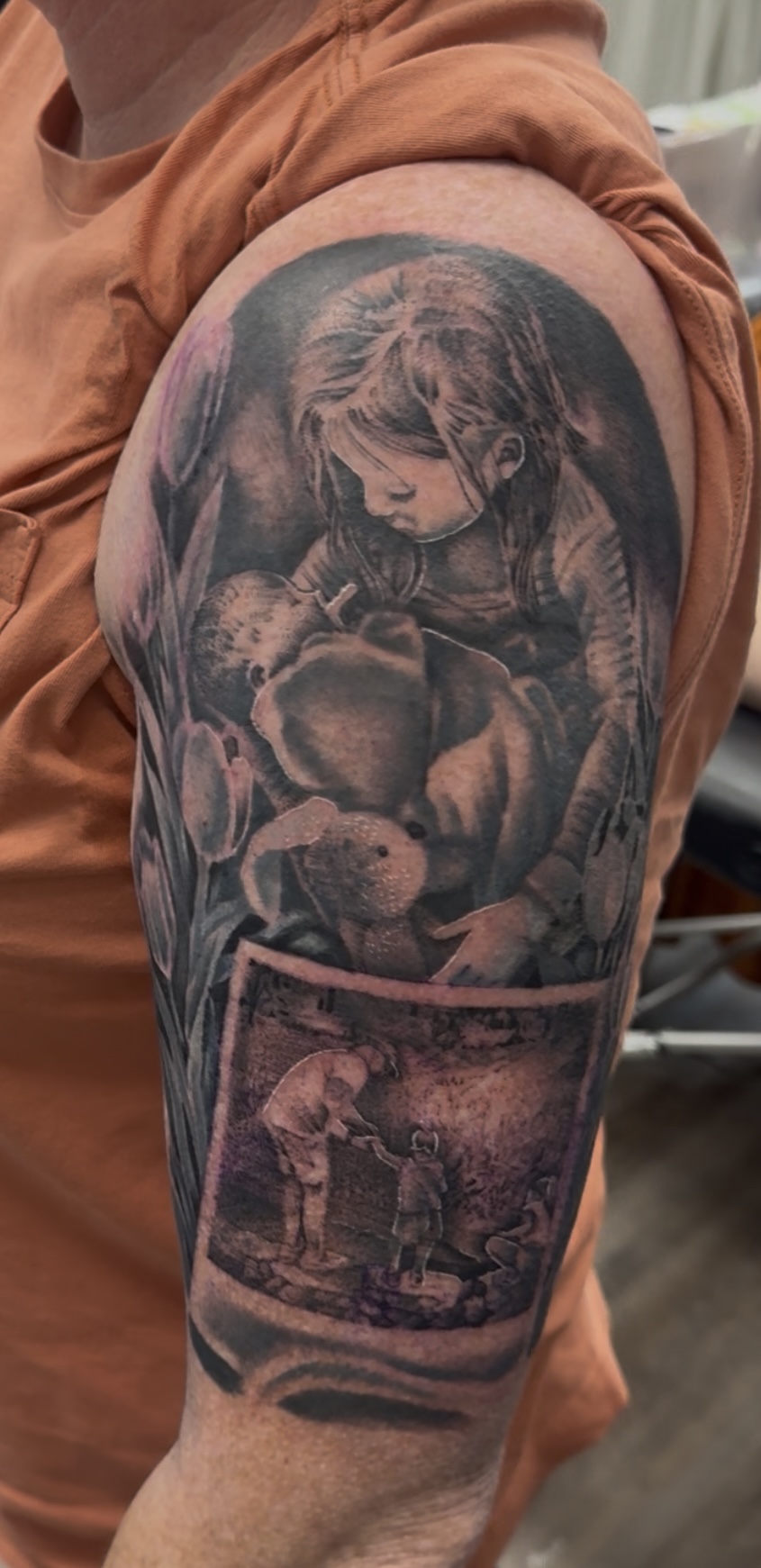 Tattoo of a girl with a teddy bear on upper arm, with a landscape scene below, in black and gray ink.