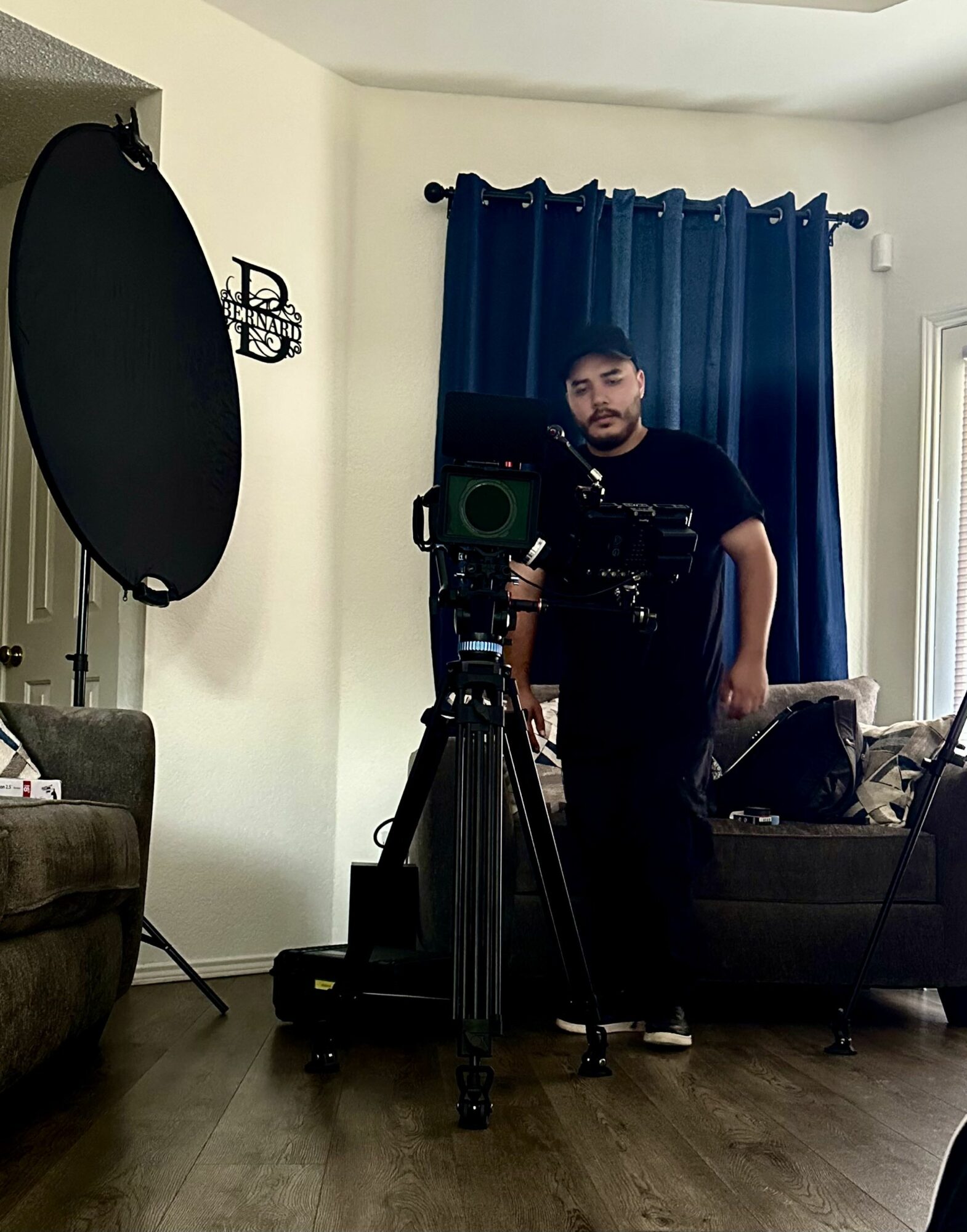 Man taking a selfie in front of a camera on a tripod in a living room with blue curtains and furniture.