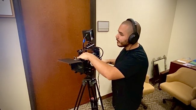Man with headphones operating camera equipment indoors, door and furniture visible in background.
