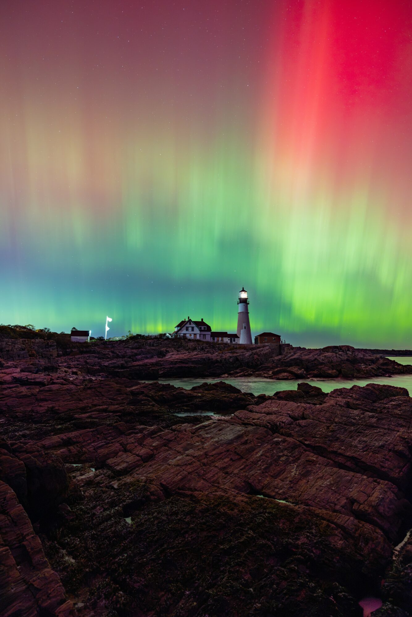 Lighthouse on rocky coast with colorful aurora borealis in the sky above, vibrant green and red lights, night scene