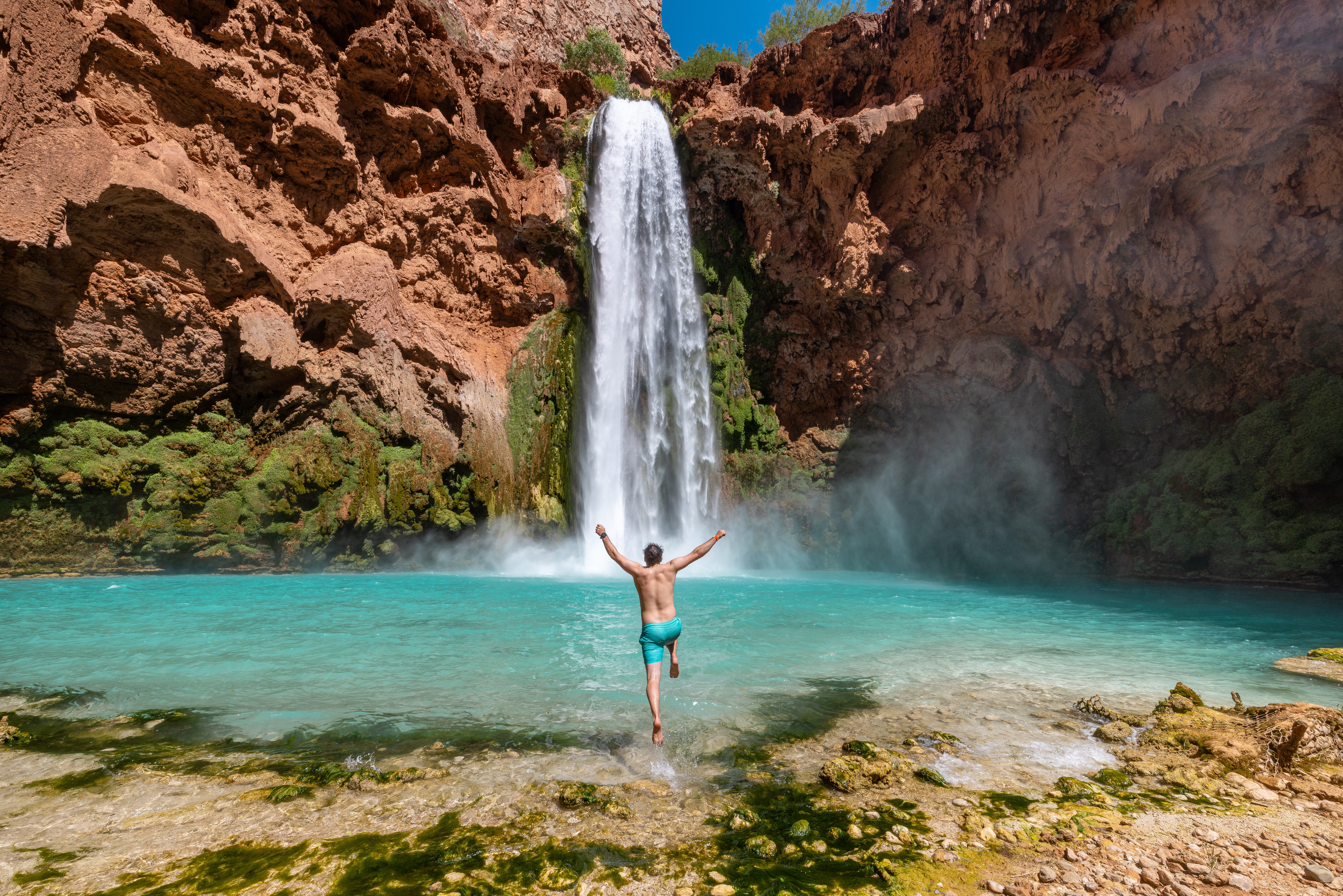 Person standing in water with arms raised in front of a waterfall and rocky canyon, turquoise water, and blue sky.
