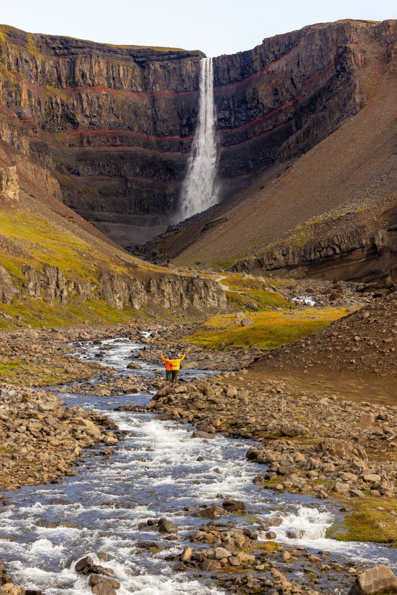 Waterfall in a canyon with a person standing in a stream, surrounded by rocky terrain and green moss, under a cloudy sky.