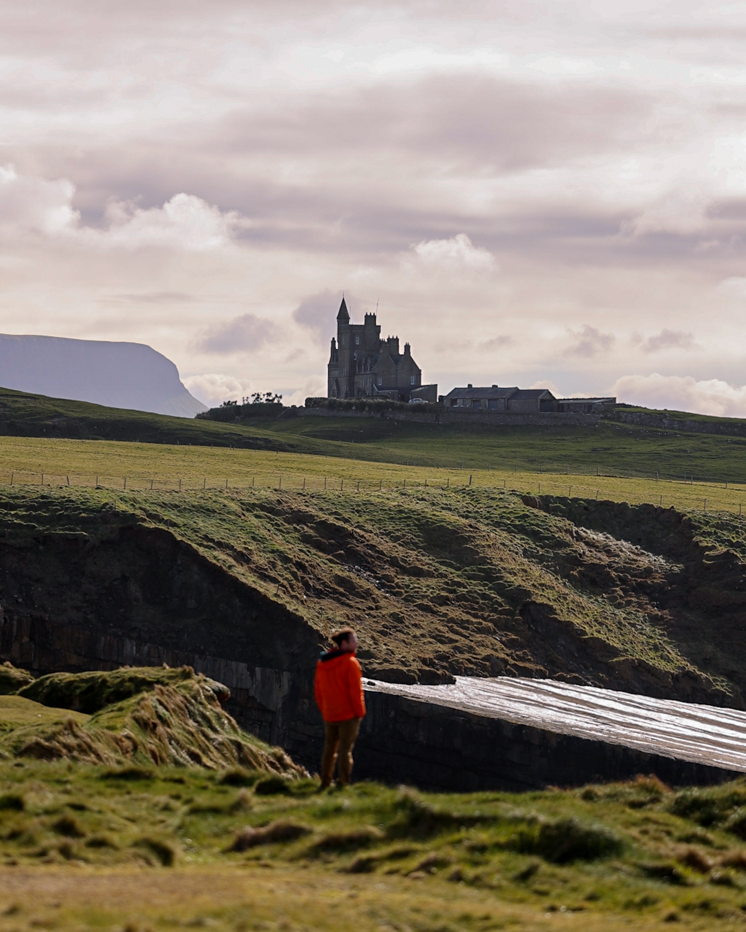 Person in red jacket standing on grassy cliff overlooking a castle on a hill under cloudy sky.
