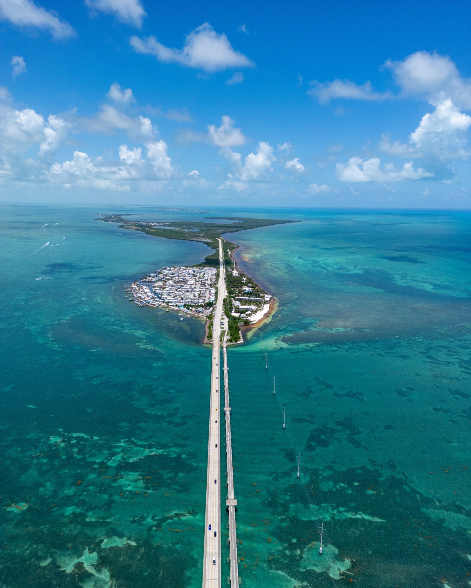 Aerial view of a bridge connecting two landmasses over turquoise water, with a blue sky and scattered clouds.
