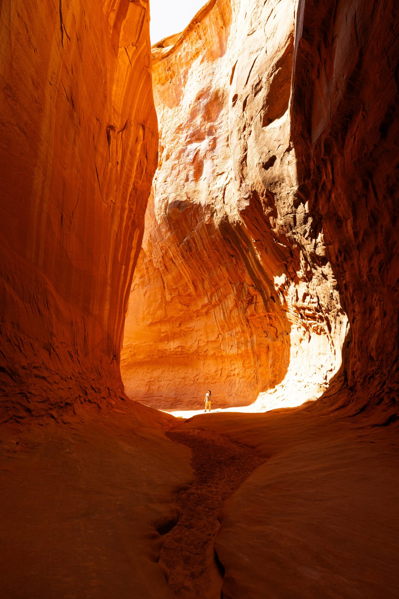 Narrow canyon with smooth, orange and white rock walls, sunlight illuminating the opening, person standing in distance.