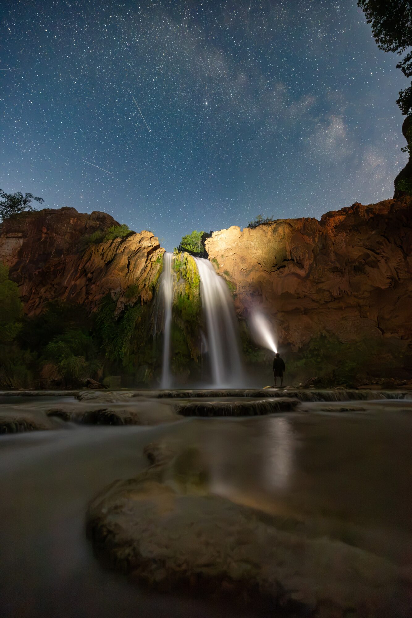 Night sky with stars above a waterfall and a person standing nearby, illuminated by a flashlight, with trees on the right.