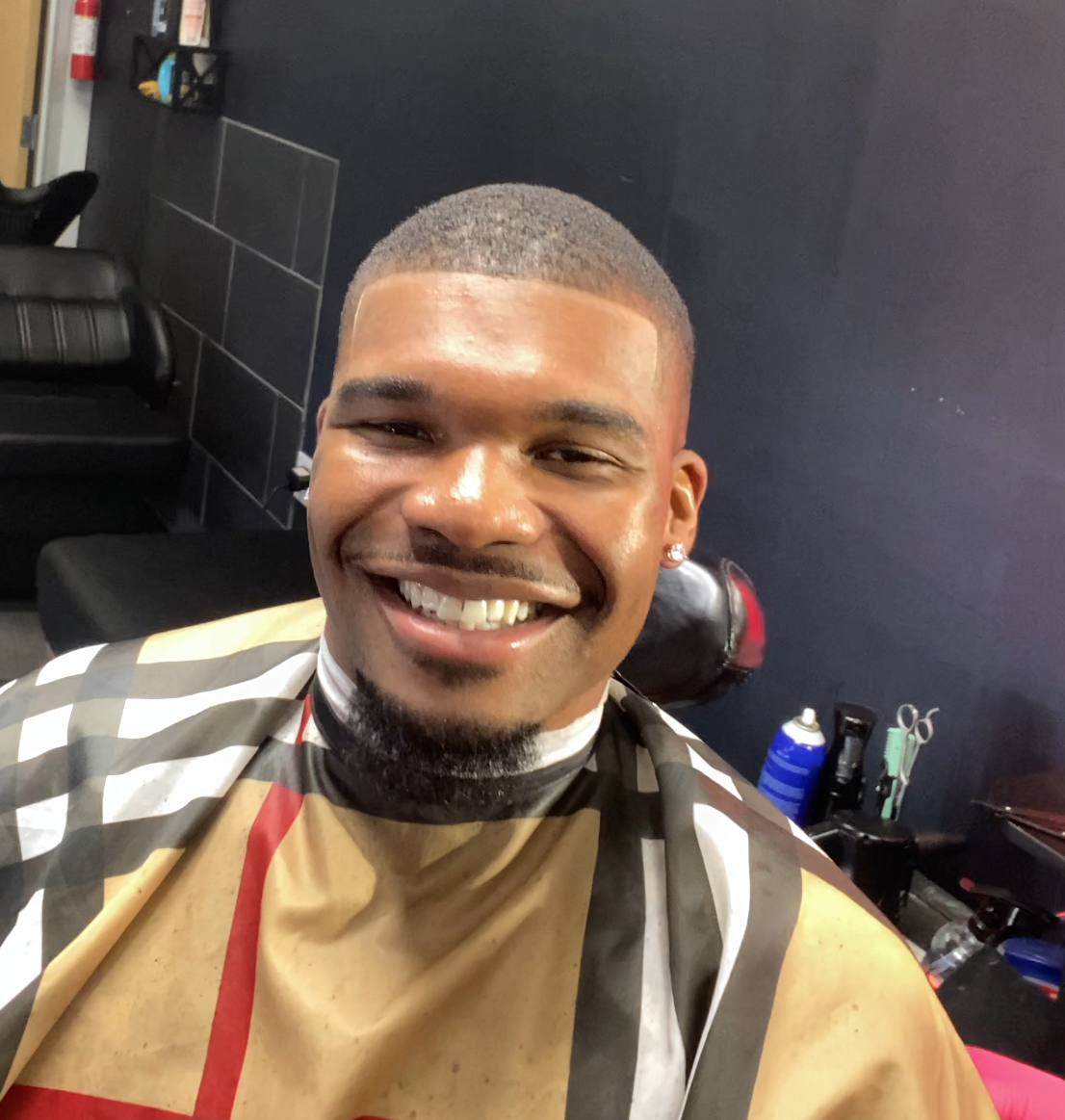 Smiling man with short hair and earrings, sitting in a barbershop chair, wearing a barber cape, with black tiled wall behind him.