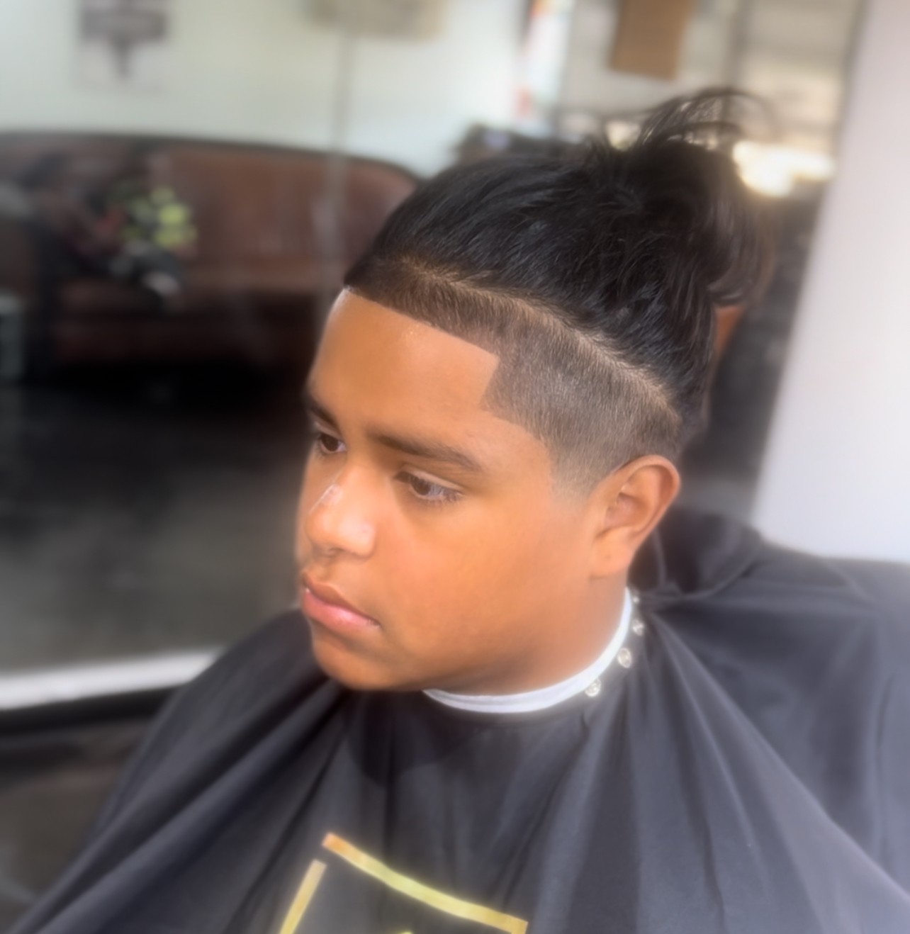 Young person with a high fade haircut and long hair tied back, sitting in a salon chair, wearing a black cape.